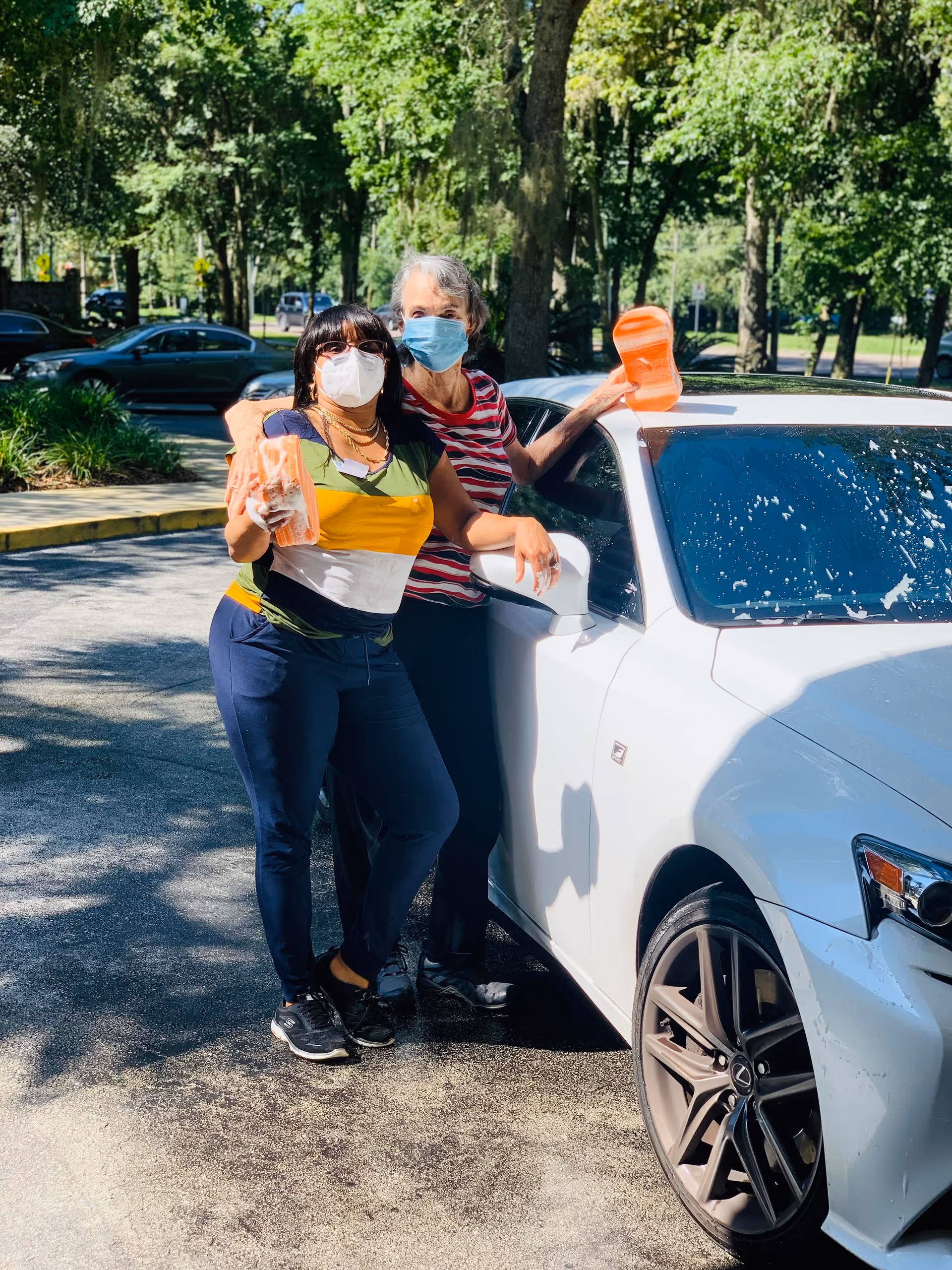 Two women wearing face masks washing a white car outdoors on a sunny day. One woman is holding an orange sponge and the other is leaning on the car with soap suds visible on the car's windshield and hood. Trees and parked cars are visible in the background.