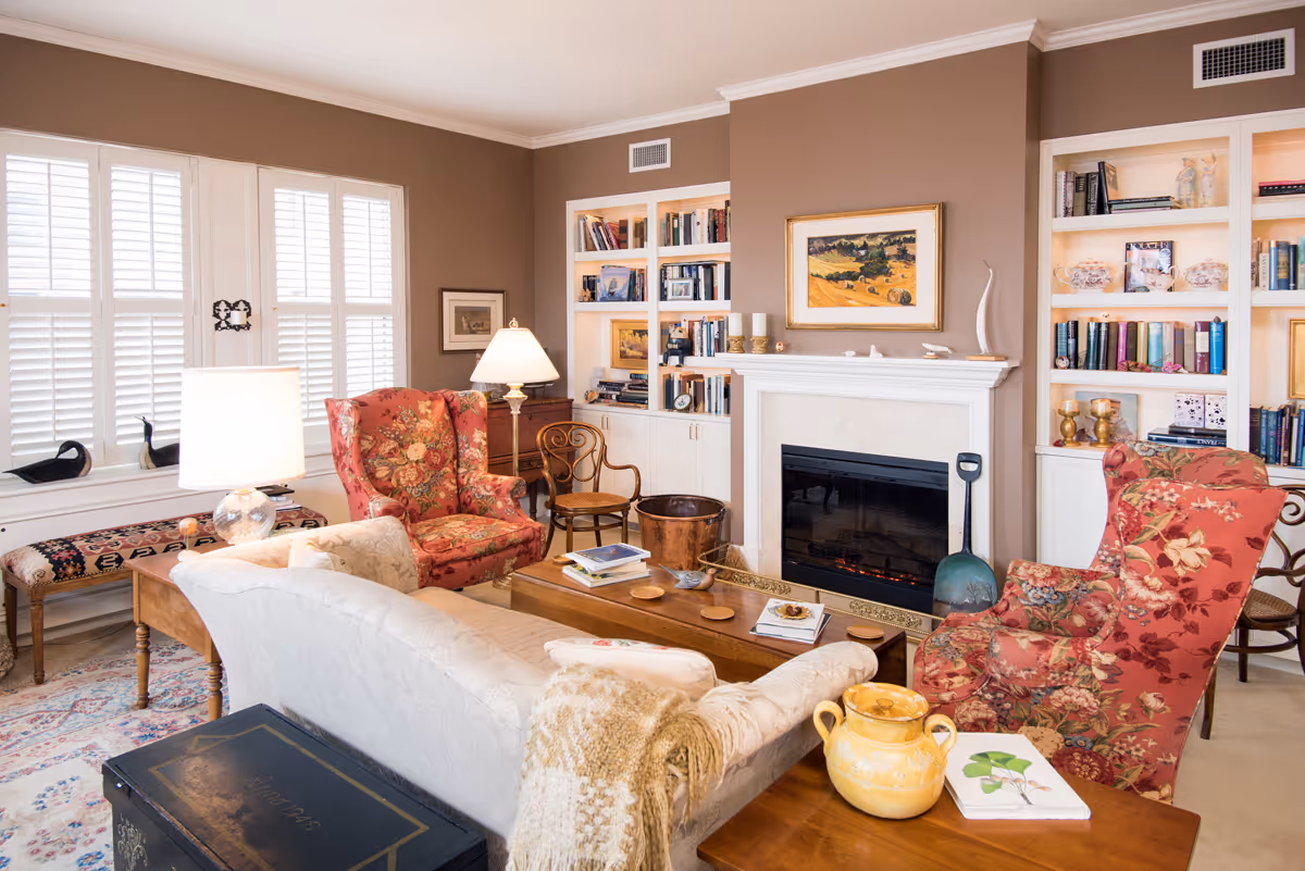 A cozy living room with beige walls and white trim, featuring a white couch with a beige throw blanket, two red floral armchairs, a wooden coffee table with books and coasters, a fireplace with a painting above it, built-in white bookshelves filled with books and decorative items, a wooden chair, a lamp on a side table, and large windows with white shutters letting in natural light.