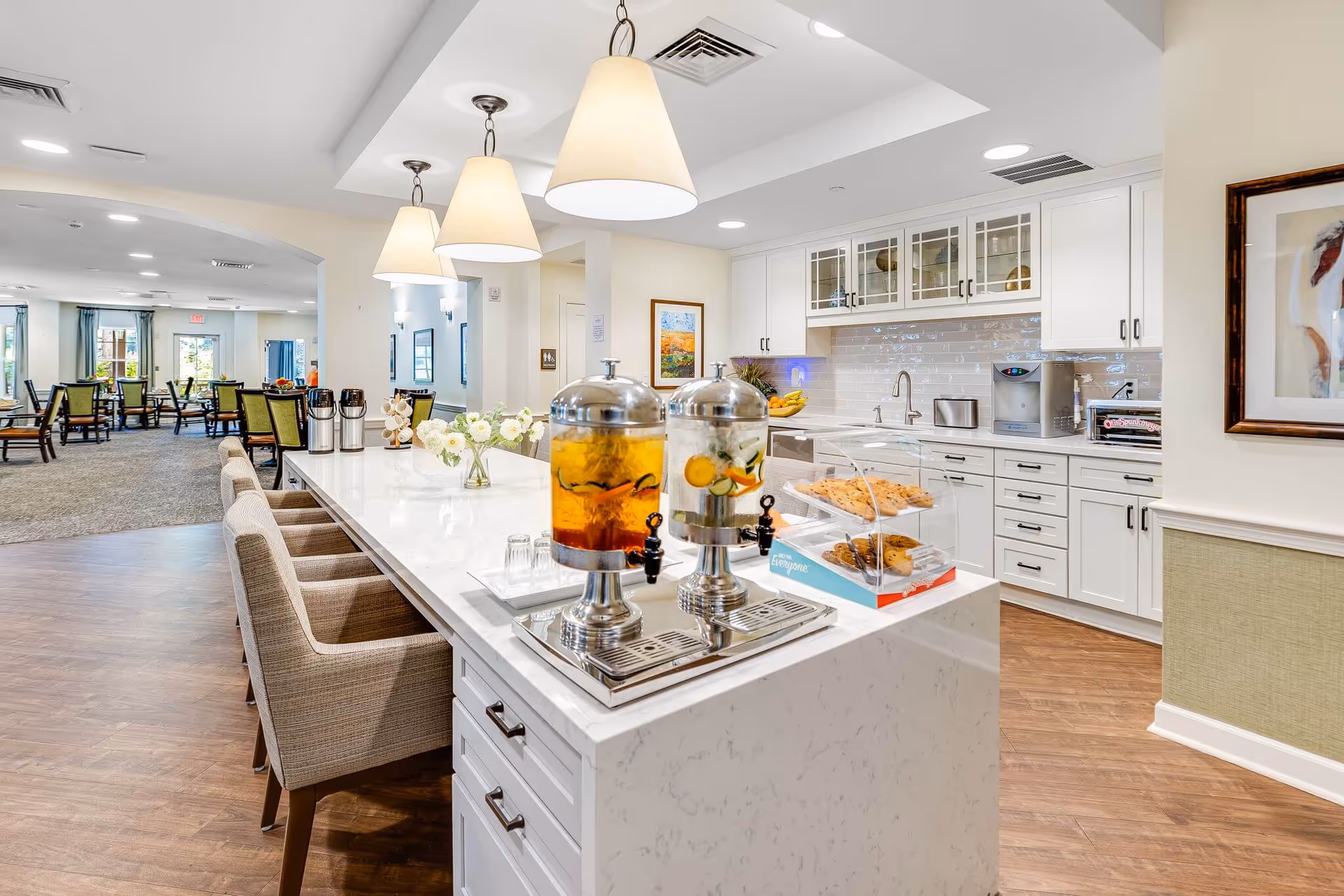A bright and modern kitchen and dining area in a senior living facility. The kitchen features white cabinetry, a white marble countertop island with beige cushioned chairs, two beverage dispensers with infused water and iced tea, a display case with baked goods, and three pendant lights hanging above the island. In the background, there is a dining room with tables and chairs, large windows with blue curtains, and framed artwork on the walls.