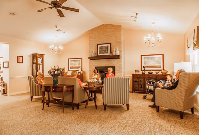 A cozy living room in a senior living facility with several elderly people seated on armchairs and sofas arranged around a coffee table. The room features a brick fireplace with decorative items on the mantel, framed artwork on the walls, a grandfather clock, and warm lighting from chandeliers. The carpeted floor and neutral color palette create a comfortable and inviting atmosphere.