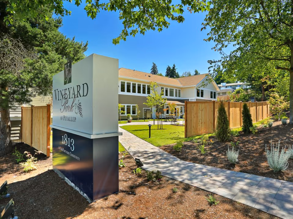 Outdoor view of Vineyard Park of Puyallup senior living facility showing a large sign with the facility name and address, a paved walkway leading to a two-story building surrounded by green lawn, trees, and a wooden fence under a clear blue sky.