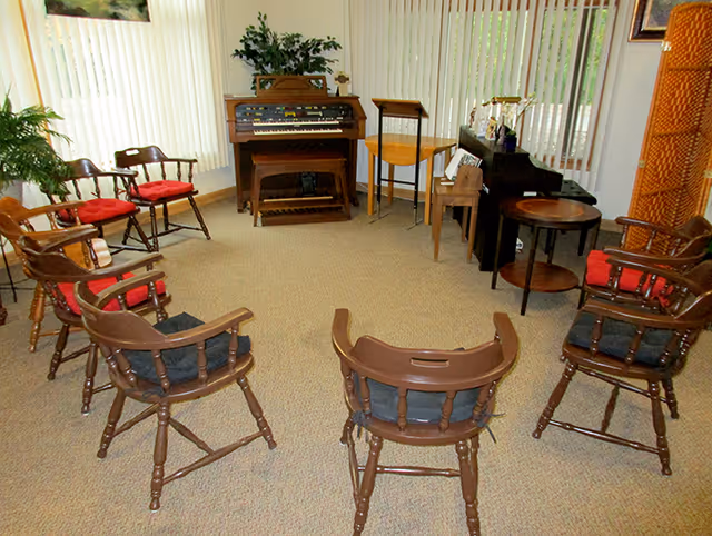 A small communal music room with wooden chairs arranged in a circle around an organ and a piano by large vertical blinds.