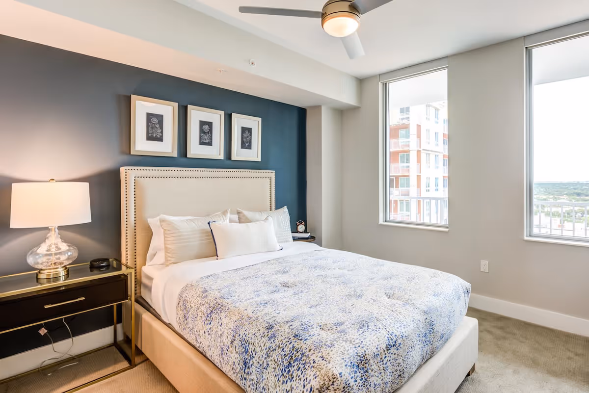 A bright bedroom with a beige upholstered bed featuring a blue and white patterned comforter, three framed botanical prints on a dark accent wall above the headboard, a glass table lamp on a black nightstand, and two large windows letting in natural light with a view of nearby buildings.