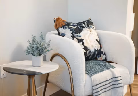 Plush white accent chair with a floral pillow and striped throw beside a small round side table topped with a potted plant.