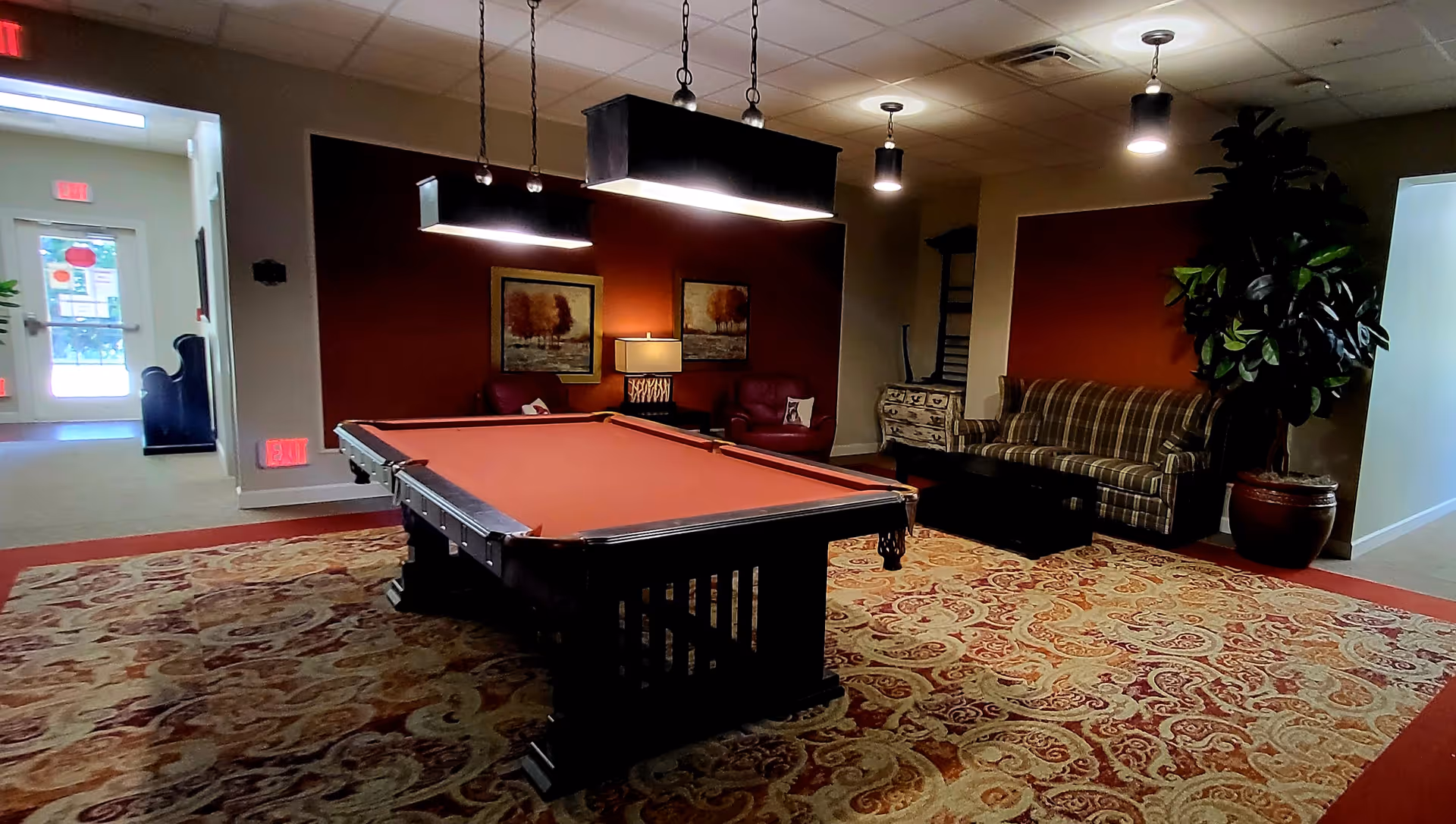 A cozy indoor recreational room featuring a pool table with a reddish felt surface in the center, illuminated by two hanging rectangular lights. The room has a patterned carpet, a plaid sofa, two red armchairs, a small table with a lamp, framed artwork on the walls, and a large potted plant. There is a hallway leading to an exit door with windows in the background.