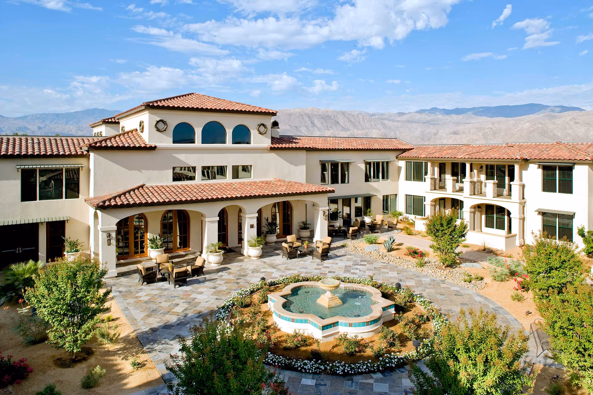 Exterior view of Segovia facility featuring a courtyard with a decorative fountain surrounded by flowers and greenery. The building has a Mediterranean architectural style with white walls, red tile roofs, arched windows, and outdoor seating areas. Mountains and a partly cloudy sky are visible in the background.