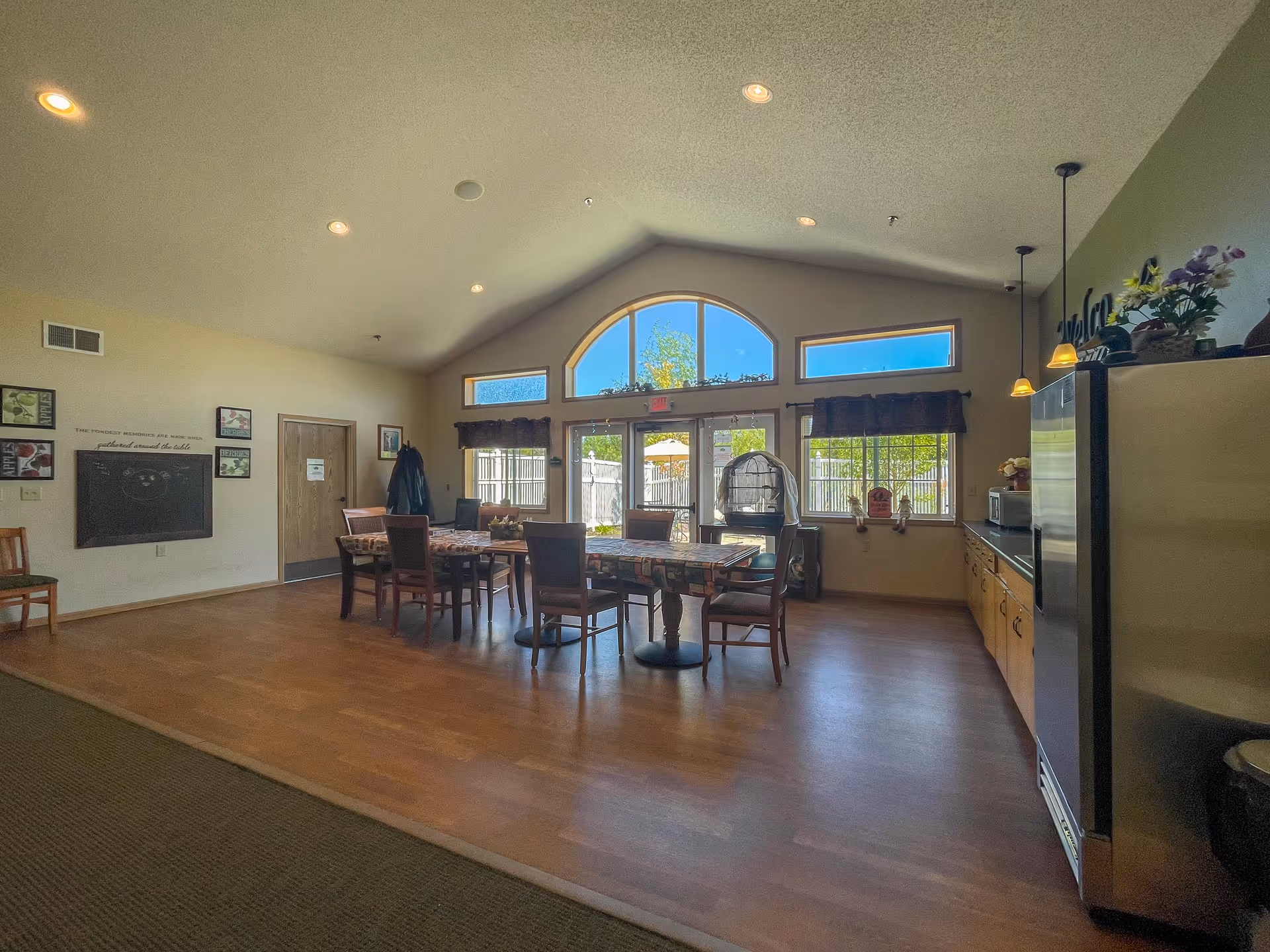 A spacious room with a high vaulted ceiling and large arched windows letting in natural light. There is a long table with several chairs around it in the center of the room. On the right side, there is a kitchen area with a refrigerator, cabinets, and hanging pendant lights. The walls are decorated with framed pictures and a chalkboard. The floor is wooden, and the room has a warm, inviting atmosphere.