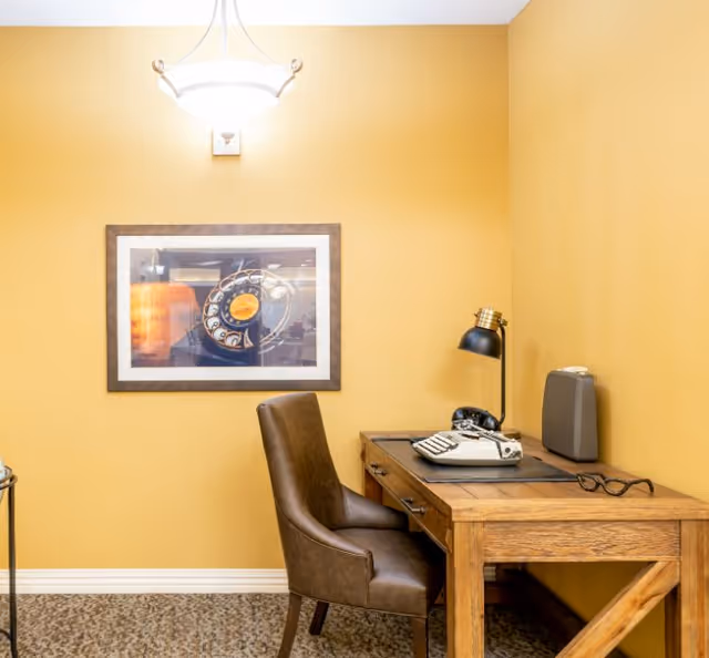 A cozy office nook with a wooden desk, leather chair, desk lamp, vintage typewriter and rotary phone against yellow walls and framed artwork.