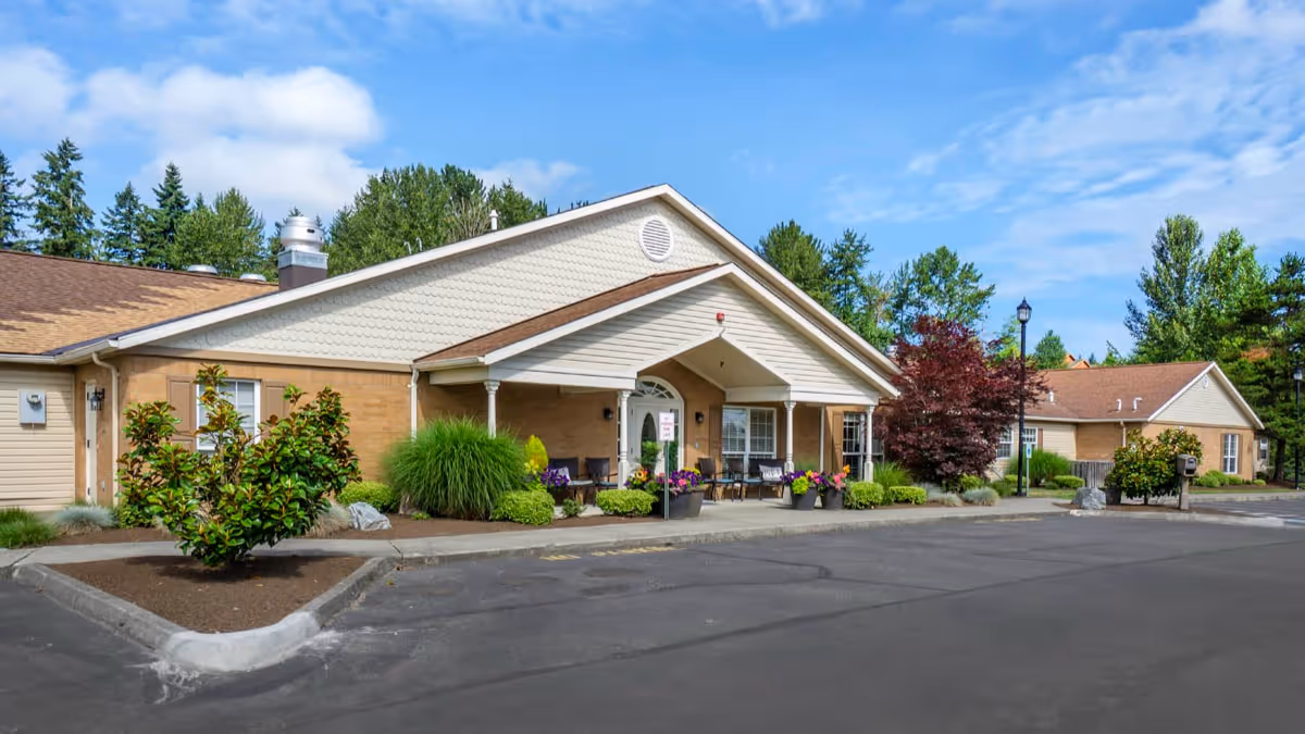 Exterior view of a single-story senior living facility building with a covered entrance, surrounded by landscaped greenery and trees under a partly cloudy sky.