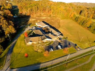 Aerial view of Jamestowne Assisted Living facility surrounded by green fields and trees with autumn foliage. The building complex includes multiple connected structures with parking spaces and a driveway leading to the main road.