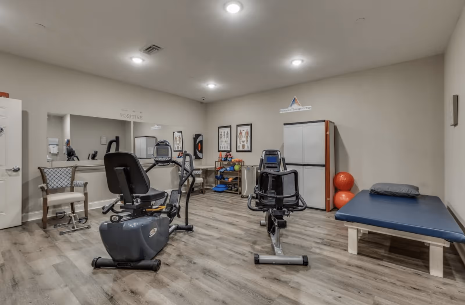 A therapy or exercise room with two stationary exercise bikes, a padded therapy table with a pillow, a chair, and a storage cabinet. The room has light-colored walls, wooden flooring, and framed anatomical charts on the wall. There are also exercise balls and various therapy equipment on a small shelf.