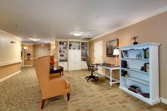 A well-lit interior hallway area in a senior living facility featuring a beige carpet with a patterned design, two tan upholstered chairs with a small round table in between, a white bookshelf filled with books, a wooden desk with a lamp and a swivel chair, and a white cabinet with shelves containing books and decorative items.