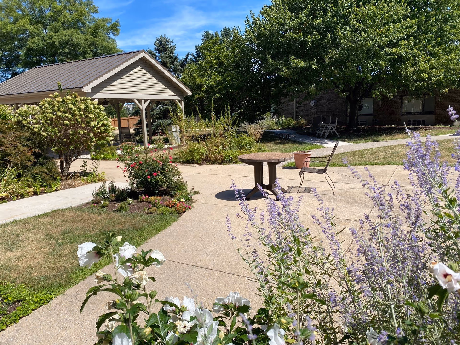 Outdoor garden area at Oak Hill Village featuring a paved walkway, a round table with a metal chair, various flowering plants including white and purple flowers, a small covered pavilion, and trees providing shade near a brick building.