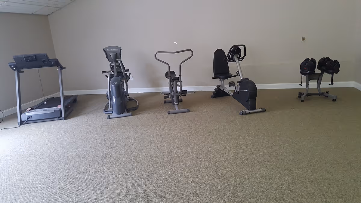 A small exercise room with a treadmill, elliptical machine, stationary bike, and a set of adjustable dumbbells on a rack against a beige wall and carpeted floor.