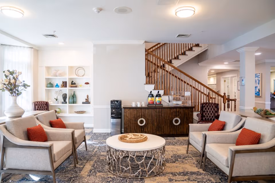 A bright and inviting senior living facility common area with beige armchairs featuring red cushions arranged around a round coffee table with a decorative centerpiece. Behind the seating area is a wooden cabinet with coffee dispensers and cups, and a staircase with wooden railings. To the left, there is a white built-in shelving unit with decorative items and a large vase with flowers near a window with sheer curtains.