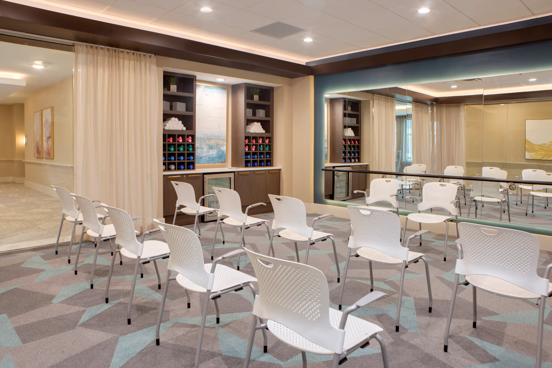 A well-lit room with multiple white chairs arranged in rows facing a mirrored wall. The room has a patterned carpet, a built-in shelving unit with colorful dumbbells and rolled towels, and a small refrigerator. The walls are painted in neutral tones with a large abstract painting visible in the reflection.