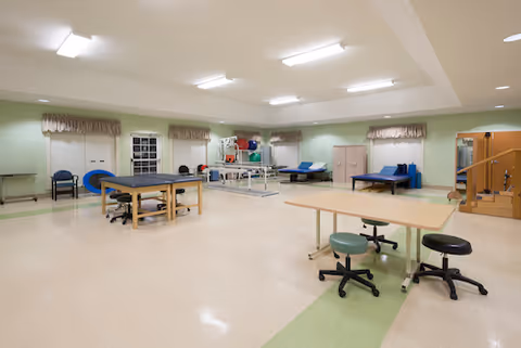 Well-lit rehabilitation room with treatment tables, exercise equipment, parallel bars, and rolling stools.