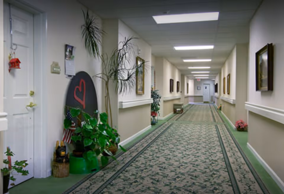 Long carpeted interior hallway of a senior living facility with plants, framed pictures, handrails and doors along the walls.
