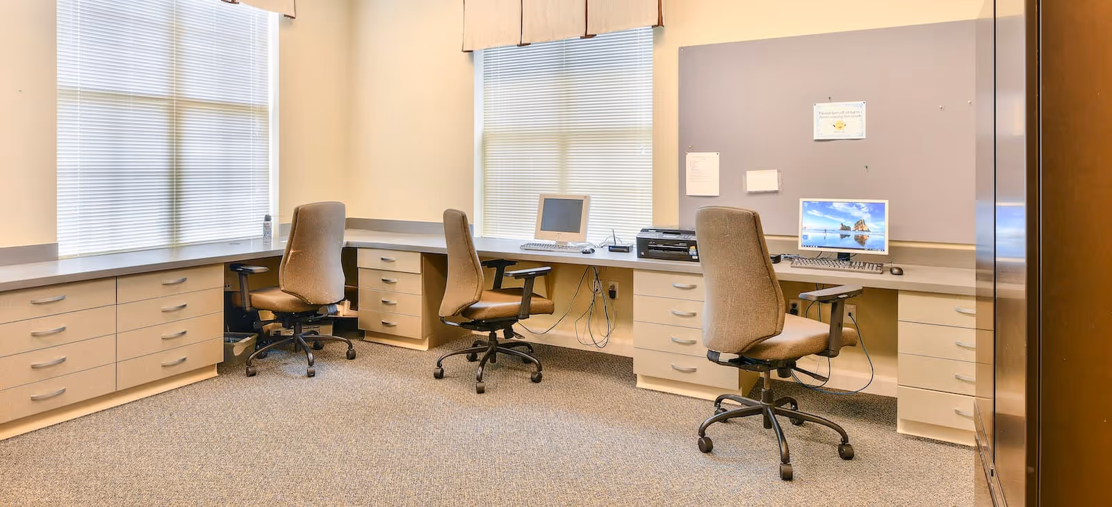 A well-lit office area with three brown swivel chairs positioned in front of a long desk with multiple drawers. Two computer monitors and a printer are on the desk. The room has beige walls, carpeted floor, and two large windows with blinds partially closed.