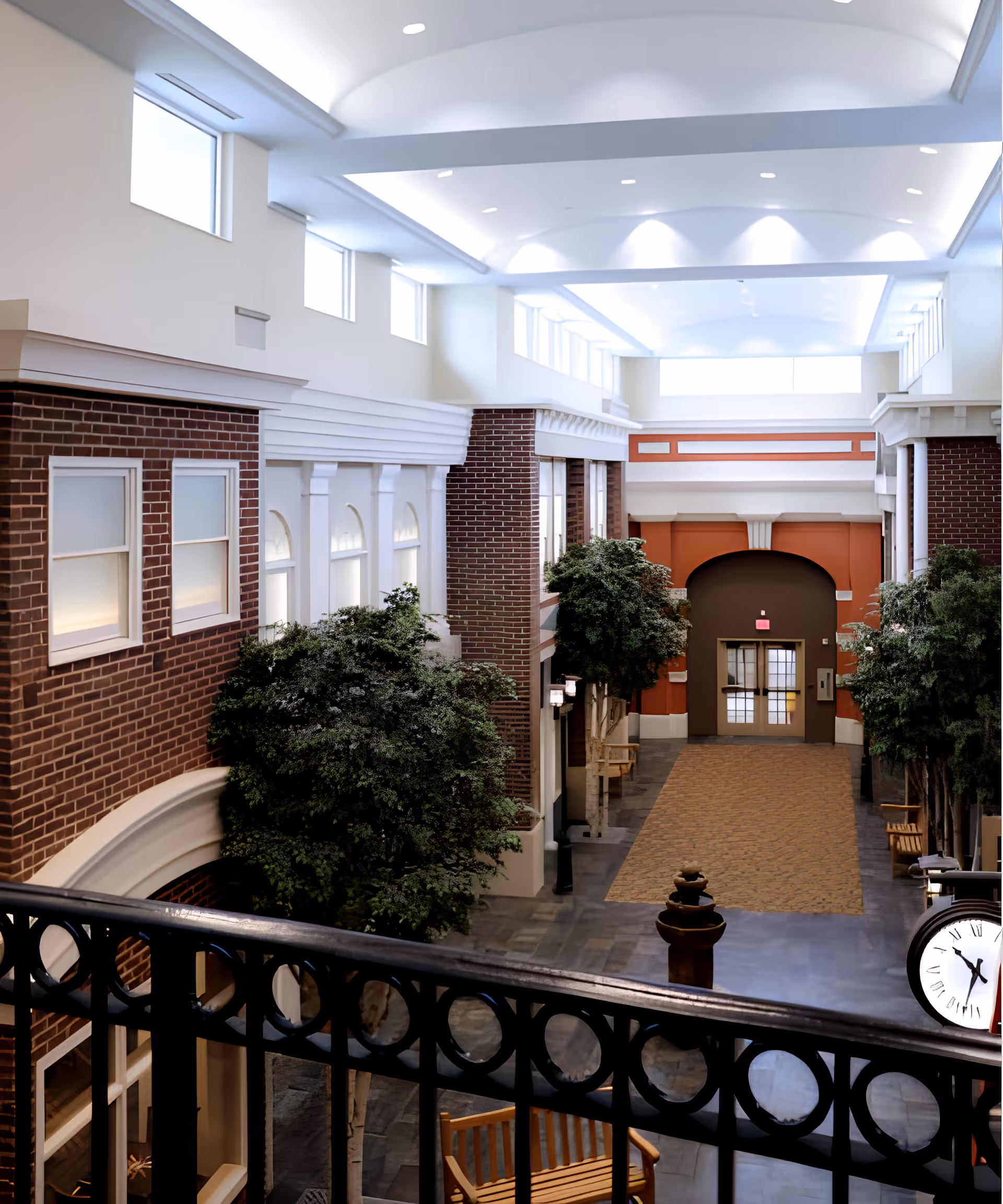 Indoor hallway of a senior living facility with high ceilings, large windows near the ceiling, brick and white walls, several green indoor trees, wooden benches, a decorative fountain, and a large clock on the right side.