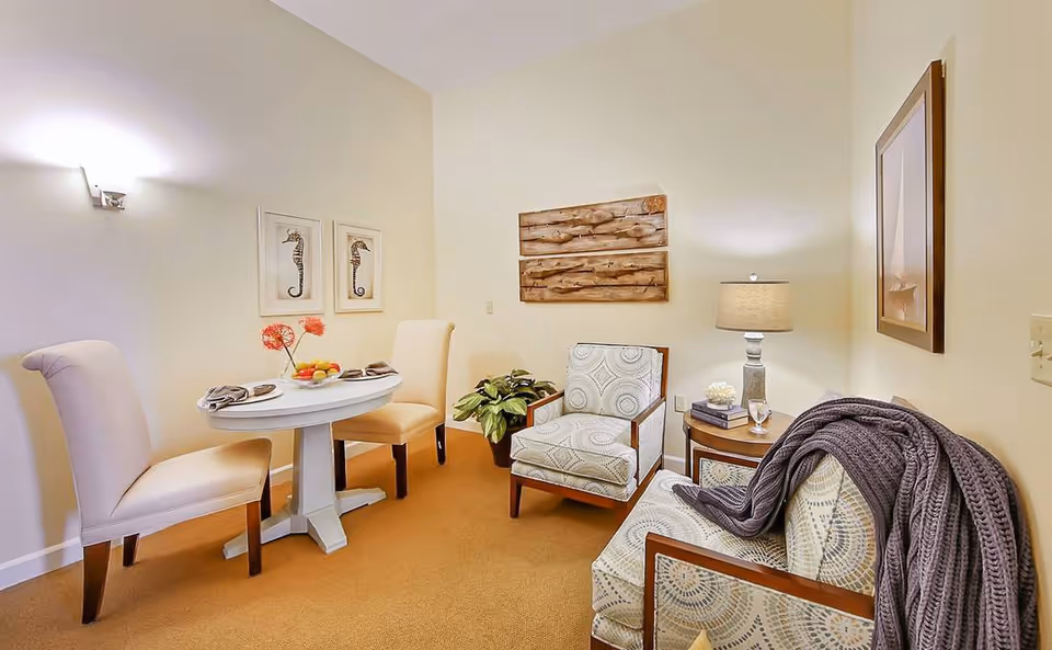 A cozy interior room with a small round white table set for two with beige upholstered chairs. There are two framed seahorse pictures on the wall above the table. Across from the table are two patterned armchairs with wooden frames, one draped with a gray knitted throw. A small round side table with a lamp, books, and decorative items is placed between the chairs. The walls are light-colored and the carpet is a warm brown tone.