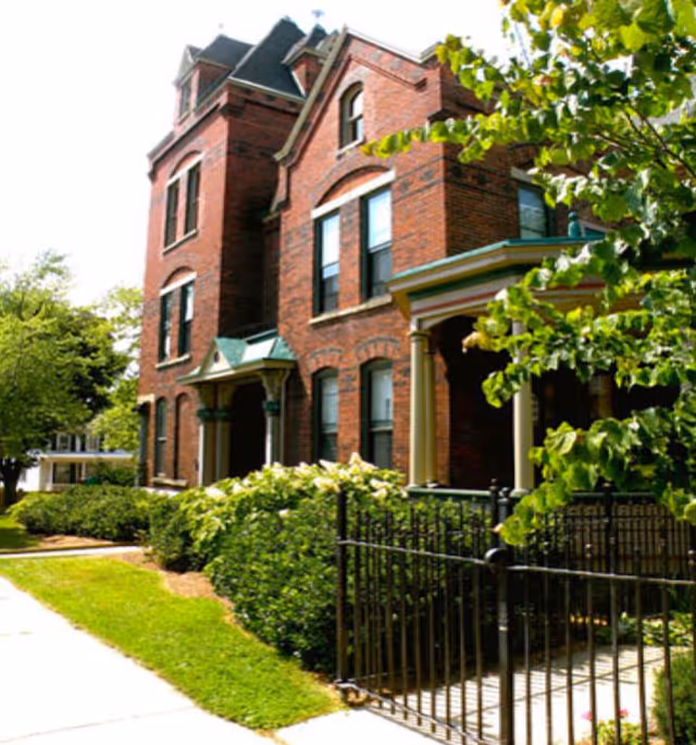 A large, historic red brick building with multiple windows and a green roof, surrounded by green bushes and trees. There is a black metal fence in the foreground and a sidewalk running alongside the building.