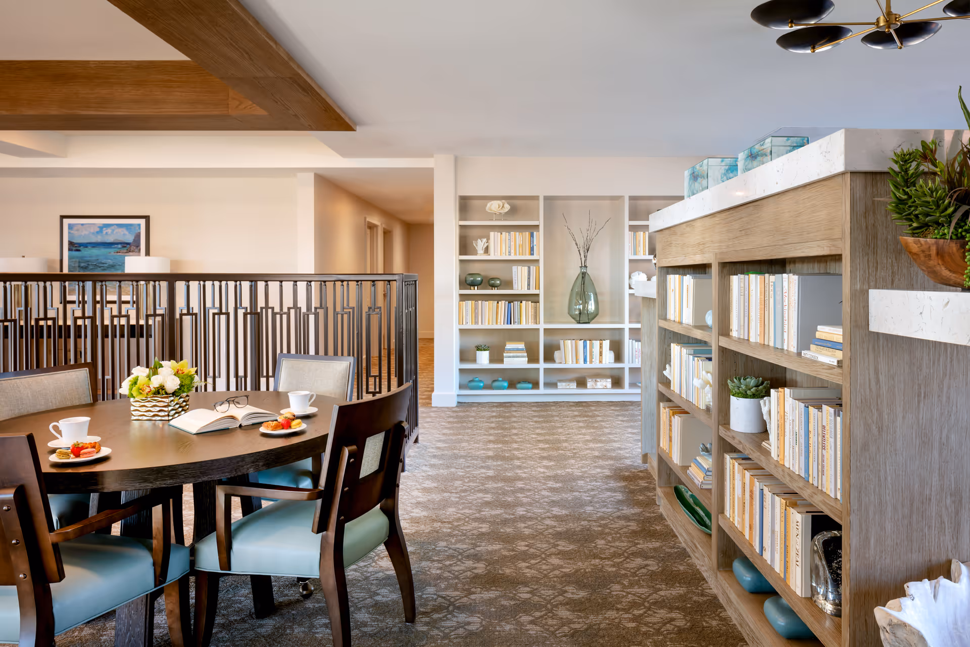 A bright and spacious interior area with a round wooden table set with cups, plates of food, and an open book. Surrounding the table are chairs with light blue cushions. To the right, there is a wooden bookshelf filled with books and decorative plants. In the background, there is a white built-in bookshelf with books and decorative items, and a hallway leading to other rooms. The space features a patterned carpet and a ceiling light fixture.