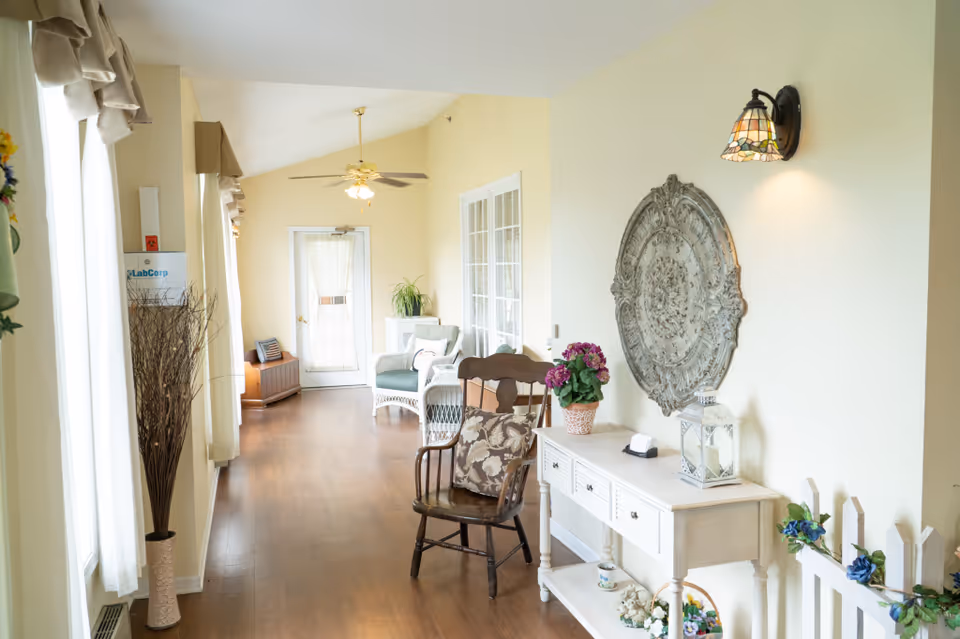 A bright hallway in a senior living facility with wooden flooring, cream-colored walls, and large windows with sheer curtains. The hallway features a wooden chair with a patterned cushion, a white console table decorated with a potted plant, a lantern, and small decorative items. A large ornate wall decoration hangs above the console table, and a Tiffany-style wall sconce provides light. In the background, there is a white wicker chair with a cushion, a ceiling fan with lights, and a door with a sheer curtain.