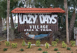 Wooden sign with a red tiled roof that reads 'LAZY DAYS VILLAGE 2524 N. TRAIL' with a painted fish decoration below the text, surrounded by trees and small plants.