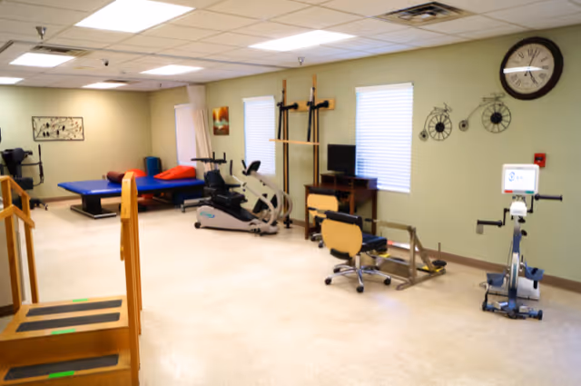 A bright rehabilitation room with stationary bikes, treatment tables, parallel bars, and other therapy equipment.