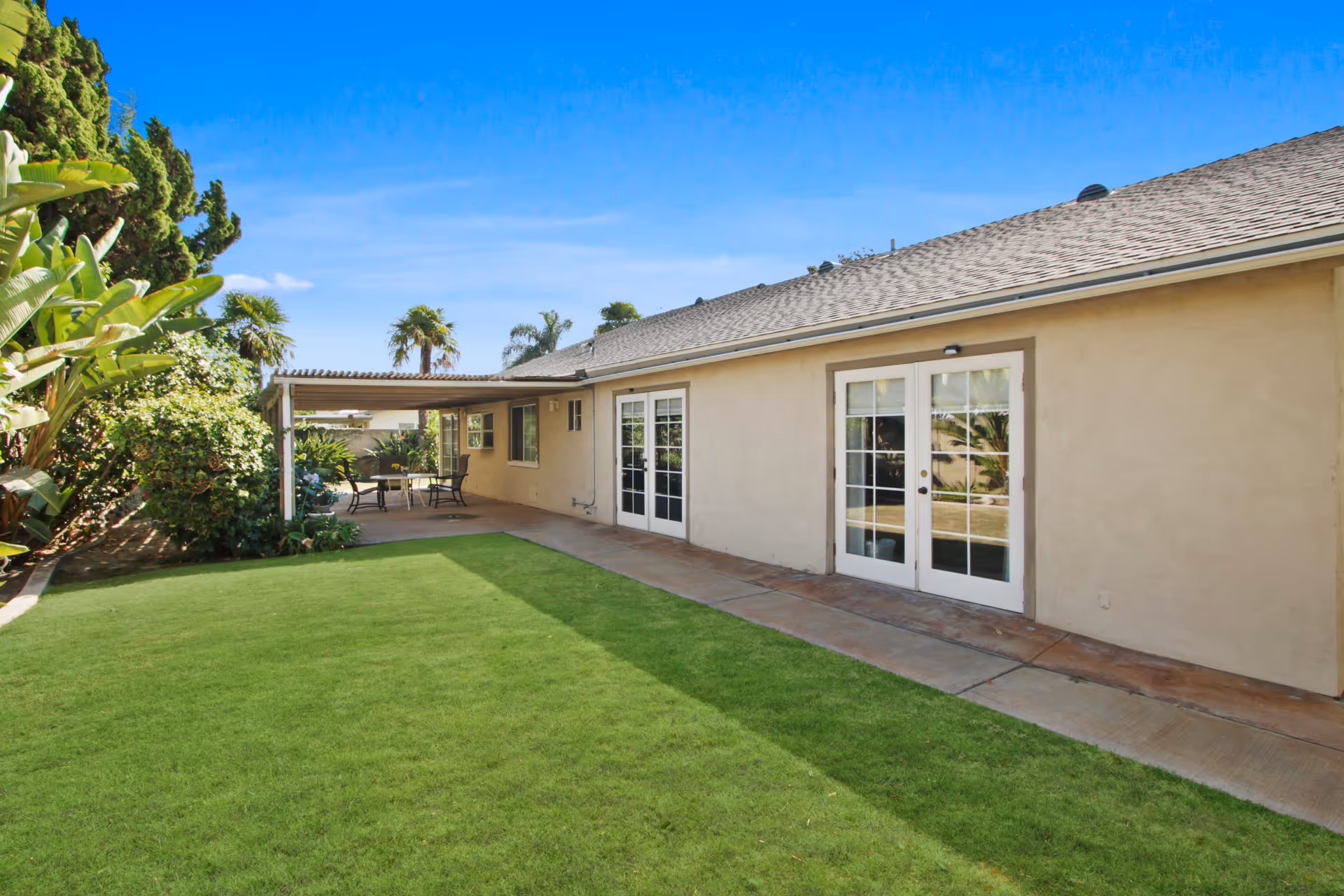 Outdoor view of a single-story building with beige walls and a shingled roof. There are two sets of white French doors leading to a covered patio area with a table and chairs. The patio is surrounded by lush green grass and various plants and trees under a clear blue sky.