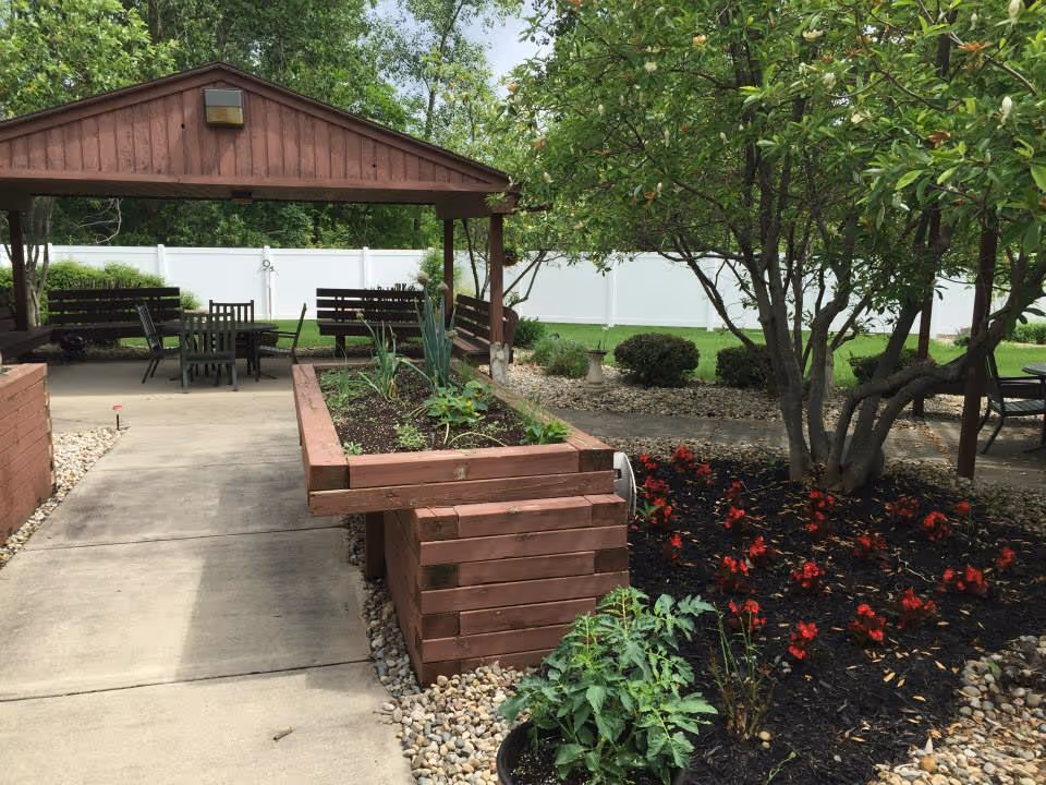 Outdoor garden area with a wooden raised planter box filled with plants, a paved walkway, a covered seating area with tables and chairs, trees, and flower beds with red flowers. A white fence and green grass are visible in the background.