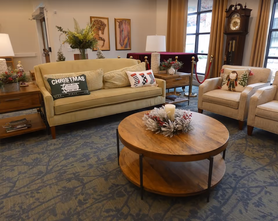 A cozy living room area with a beige sofa and two beige armchairs, each decorated with festive Christmas-themed pillows. A round wooden coffee table with a holiday wreath and candle sits in the center on a patterned carpet. Behind the sofa are two wooden side tables with lamps and holiday decorations. Large windows with beige curtains and a tall grandfather clock are visible in the background.