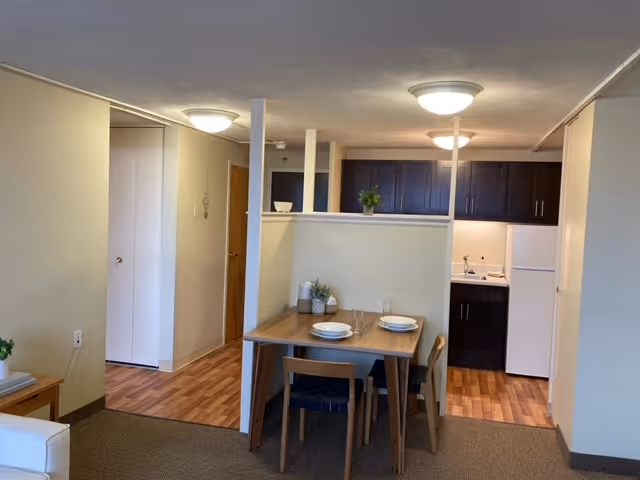 Interior view of a senior living facility unit at Golden West showing a small dining area with a wooden table set for two, adjacent to a compact kitchen with dark cabinets, a white refrigerator, and a sink. The space has beige walls, ceiling lights, and wood-patterned flooring in the kitchen and hallway area.