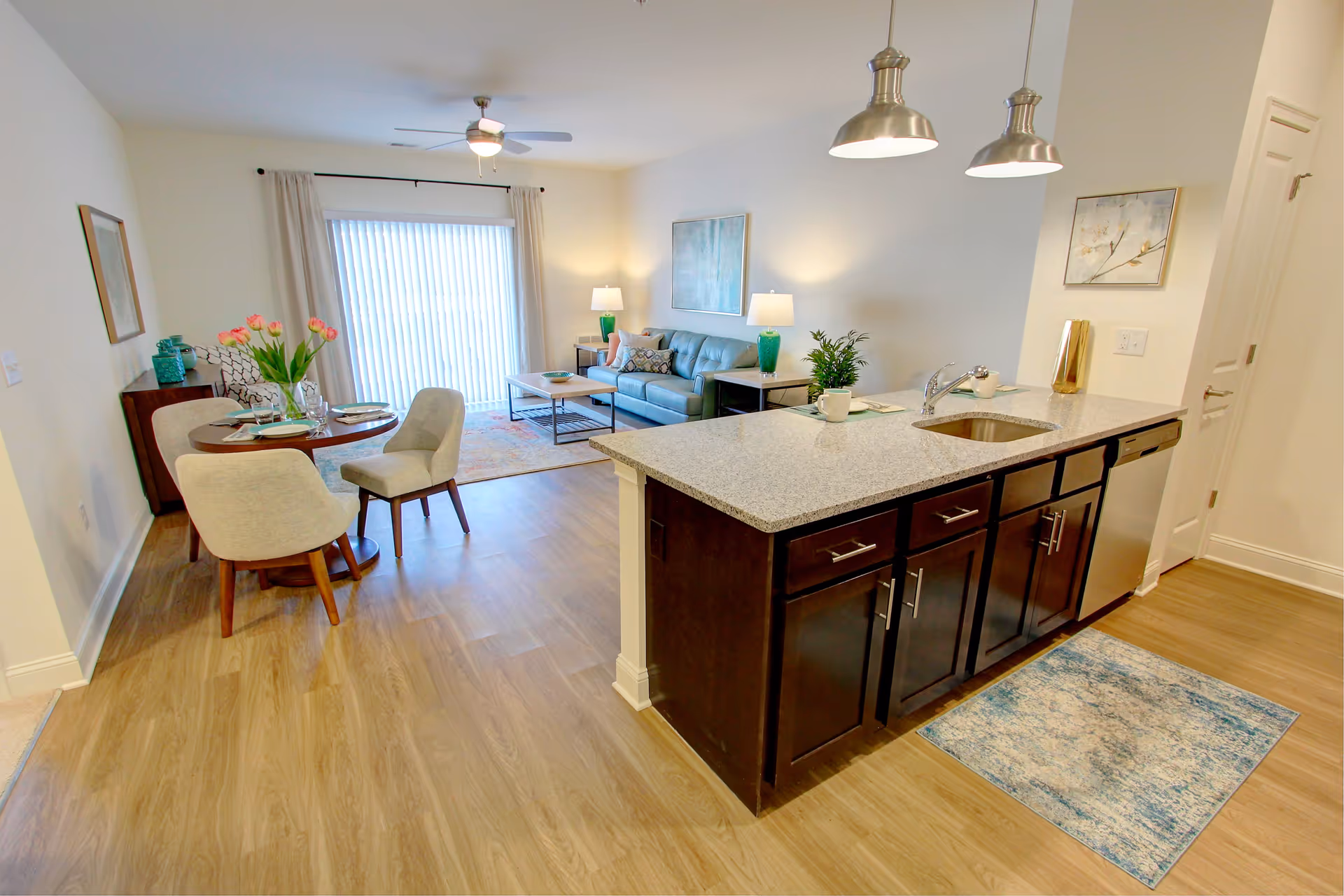 Open-concept senior apartment interior featuring a kitchen island with granite countertop and sink, a small dining area with a round table and four chairs, and a living room with a blue sofa, coffee table, two side tables with lamps, and a large window with vertical blinds.