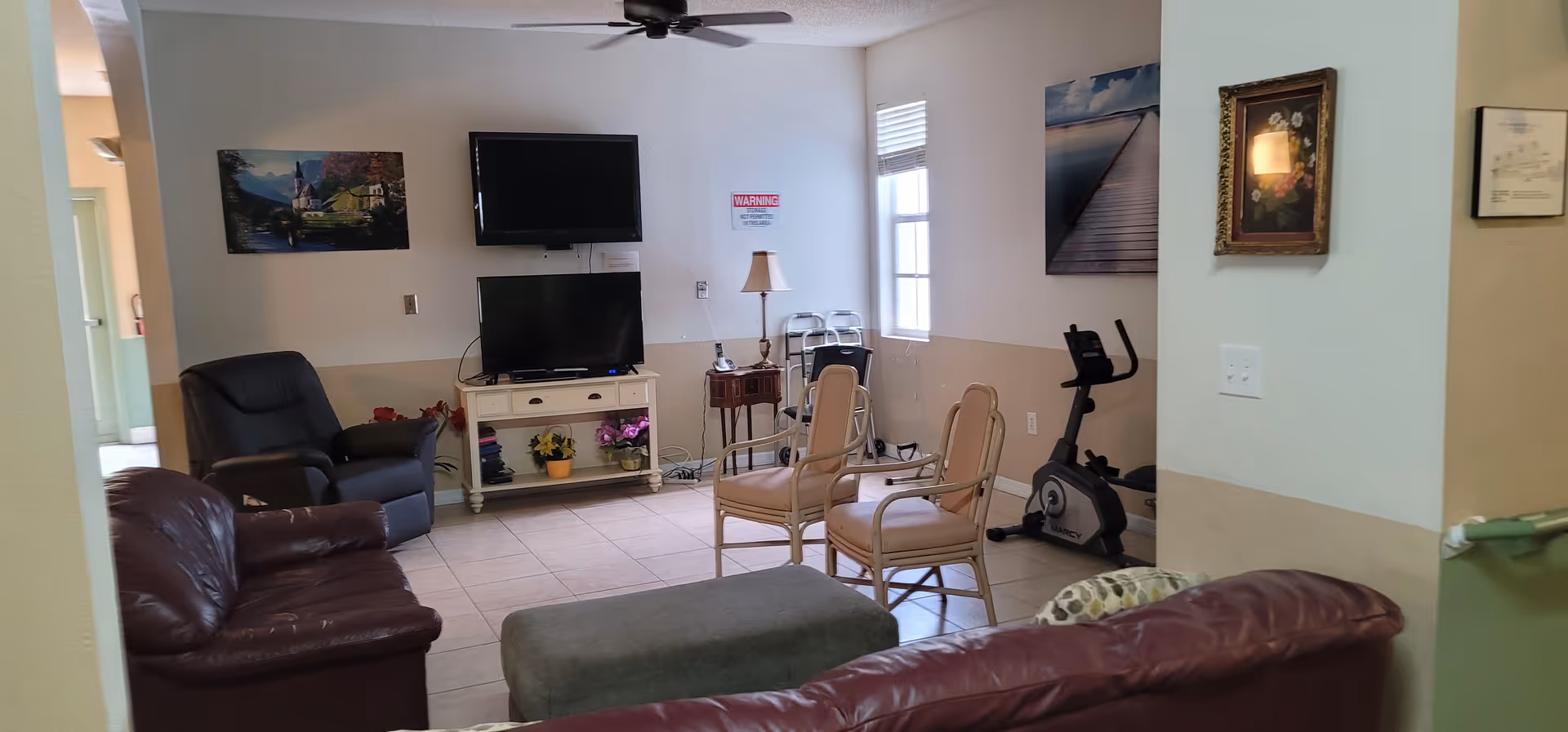 A cozy living room area with multiple seating options including a brown leather sofa, a dark recliner, and two beige chairs. There is a TV mounted on the wall above another TV on a white stand with decorative plants. The room has tiled flooring, a ceiling fan, framed pictures on the walls, a small table with a lamp, and an exercise bike near the window.