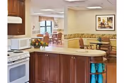 Interior view of a senior living facility showing a kitchen area with wooden cabinets, a stove, and a microwave. In the background, there is a dining area with tables and chairs, a window with curtains, and framed artwork on the wall.