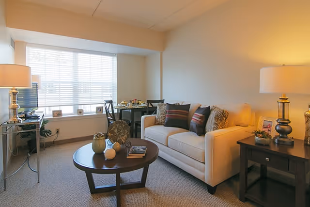 A cozy living room with a beige sofa adorned with patterned cushions, a round wooden coffee table with decorative items and books, a side table with a lamp and small plants, a dining table with chairs near a large window with blinds, and a TV on a stand in the corner.
