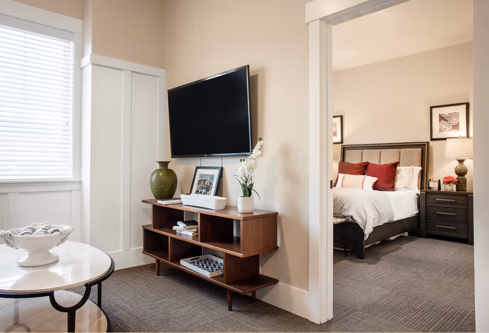A cozy living room area with a wall-mounted flat-screen TV above a wooden media console holding decorative items including a green vase, a framed photo, and a white orchid plant. To the right, an open doorway reveals a bedroom with a neatly made bed featuring white bedding and red accent pillows, a dark wooden nightstand with a lamp, and framed artwork on the walls. A round marble coffee table with a decorative bowl is partially visible in the foreground.