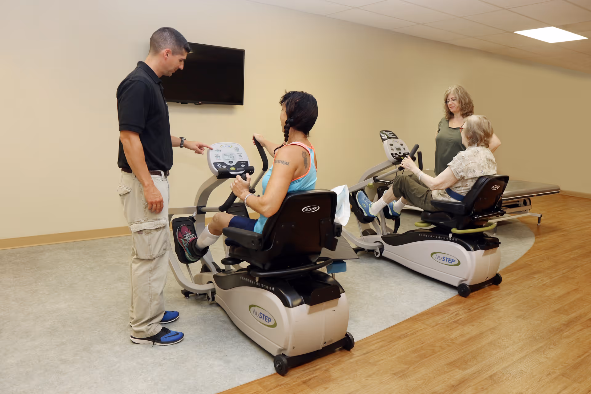 A man in a black shirt and beige pants assists a woman on a recumbent exercise bike in a fitness room. Another woman is seated on a similar exercise bike, while a third woman stands nearby watching. The room has a light-colored wall, a mounted TV, and a combination of carpet and wood flooring.
