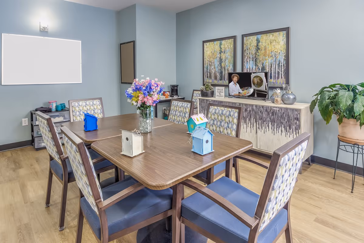 A well-lit room with a wooden table surrounded by six chairs with patterned backs and blue seats. On the table are small decorative birdhouses and a vase with colorful flowers. Against the wall is a sideboard with two framed paintings of trees, a TV displaying a painting show, and decorative items including a small plant and vases. A large potted plant is on a stand to the right, and a whiteboard is mounted on the left wall.