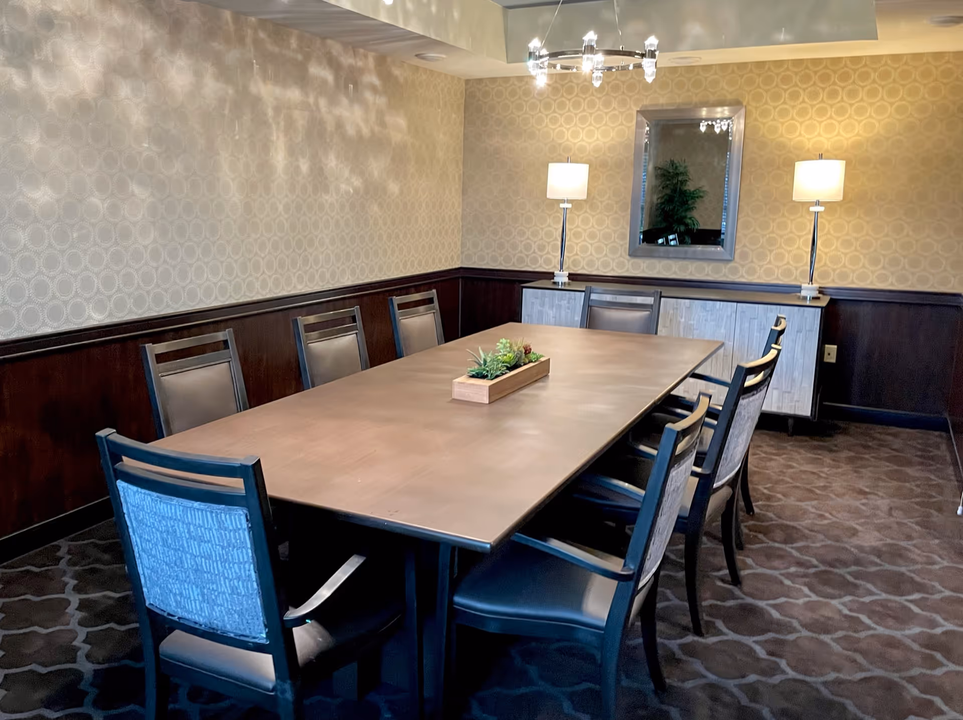 A dining room with a long rectangular wooden table surrounded by eight chairs. The room has patterned wallpaper, a carpeted floor, two table lamps on a sideboard against the wall, and a mirror hanging above the sideboard. A small rectangular planter with greenery is centered on the table.