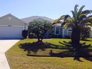 Single-story residential building with a two-car garage, a well-maintained lawn, and palm trees in the front yard under a clear blue sky.