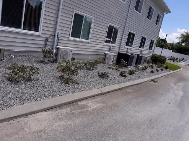 Side view of a building with beige siding, several windows, and multiple air conditioning units mounted on a gravel bed with small plants. A paved driveway runs alongside the building, and a white fence and greenery are visible in the background under a partly cloudy sky.