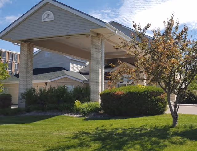 Exterior view of a senior living facility with a covered entrance supported by brick pillars, surrounded by green grass, bushes, and a tree under a partly cloudy sky.
