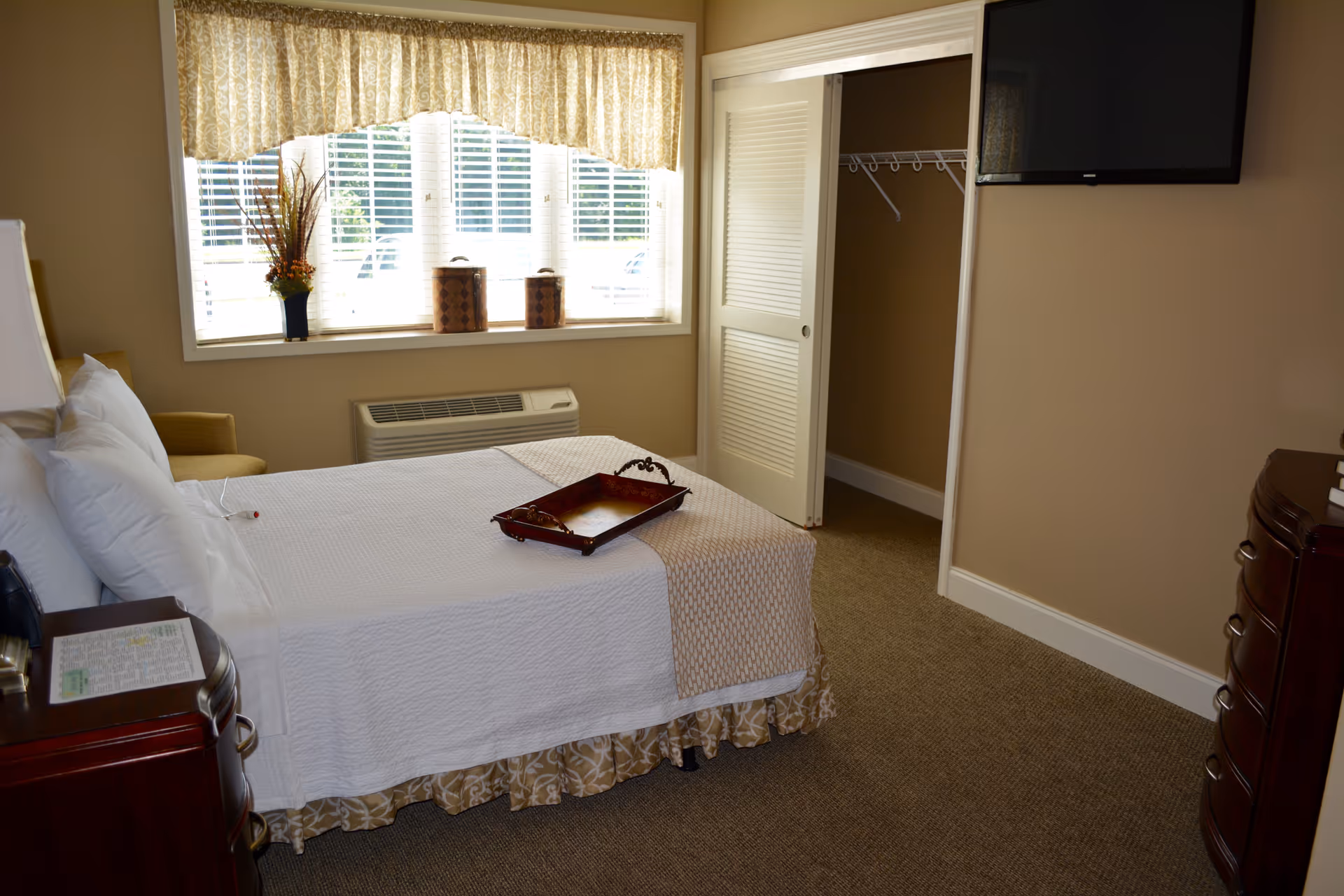 A neatly made bed with white linens and a decorative tray on top in a cozy bedroom. The room features a window with patterned curtains, a small closet with sliding doors, a wall-mounted flat-screen TV, and wooden furniture including a nightstand and dresser. The walls are painted beige and the floor is carpeted.