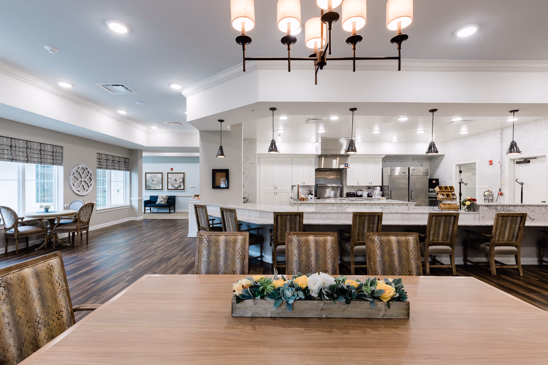 A spacious and modern dining area with a large wooden table in the foreground decorated with a rectangular planter of flowers. Behind the table are several upholstered chairs and a long counter with bar stools. The background features a bright kitchen area with white cabinetry, stainless steel appliances, and pendant lighting. To the left, there are windows with patterned shades and a small round table with chairs. The room has wood flooring and recessed ceiling lights.