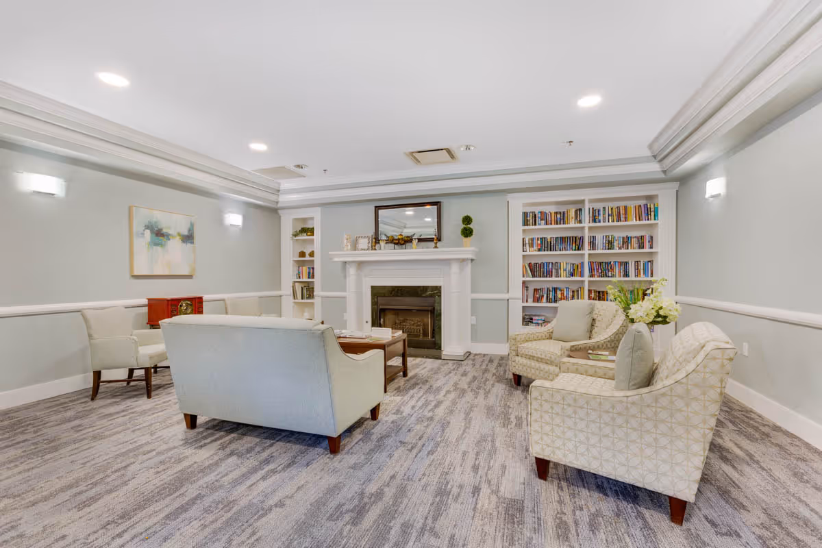 A cozy living room area with light gray walls and carpeted floor. The room features a white fireplace with a mirror above it, built-in bookshelves filled with books on either side, and several upholstered chairs and sofas arranged around a wooden coffee table. There is a piece of abstract art on one wall and soft lighting from wall sconces and ceiling lights.