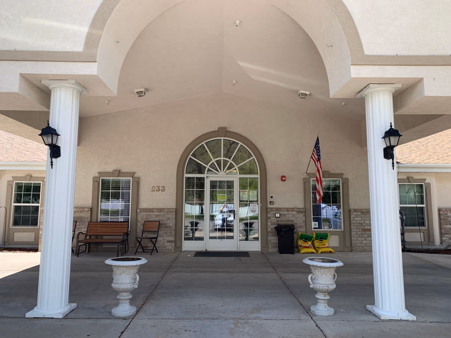 Entrance of a building with two white columns supporting an arched roof. There are two white decorative planters on the ground, a bench and a chair to the left, and an American flag mounted on the wall to the right of the glass double doors. The building number 233 is visible on the wall.