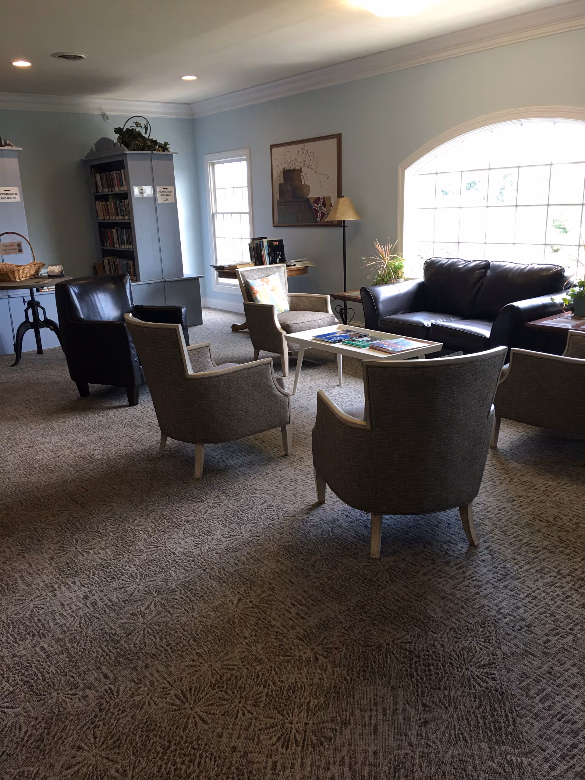 A cozy living room area with a large window letting in natural light. The room features a black leather sofa, four gray upholstered armchairs arranged around a white coffee table with books on it, a floor lamp, and a bookshelf filled with books. The walls are painted light blue and the carpet has a subtle floral pattern.